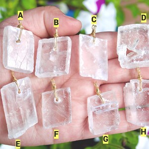 May include: Eight clear, rectangular selenite crystal pendants with gold wire wrapping. Each pendant is a different shape and size, held in a hand against a background of green and purple foliage. The pendants are translucent and have visible internal striations.
