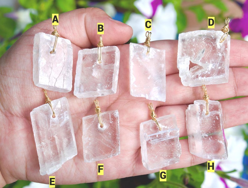 May include: Eight clear, rectangular selenite crystal pendants with gold wire wrapping. Each pendant is a different shape and size, held in a hand against a background of green and purple foliage. The pendants are translucent and have visible internal striations.
