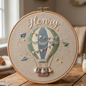 Può includere: Un telaio da ricamo circolare in legno contenente tessuto beige con una mongolfiera ricamata a mano nei toni del verde salvia, blu e crema. Il nome Henry è ricamato in corsivo bianco sopra il pallone, circondato da aeroplanini di carta e stelle.