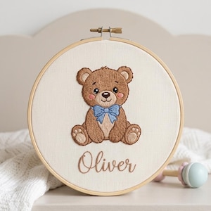 Può includere: Un telaio da ricamo circolare in legno contenente un tessuto color bianco sporco con un orsacchiotto marrone ricamato al centro. L'orso ha un papillon azzurro e guance rosa. Sotto l'orso, il nome Oliver è ricamato in corsivo marrone.