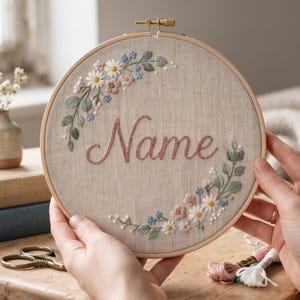 Puede incluir: Un bastidor de bordado circular de madera que contiene tela beige con la palabra Name bordada en hilo cursivo rosa. El diseño presenta una corona floral bordada con margaritas blancas, flores rosas, flores azules y hojas verdes.