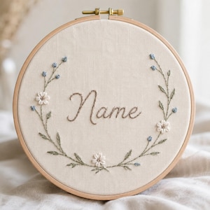 Puede incluir: Un bastidor de bordado circular de madera que enmarca una tela de color blanco roto. La tela presenta un bordado a mano de una corona floral en tonos verdes, azules y blancos, con la palabra Name centrada en letra cursiva marrón.