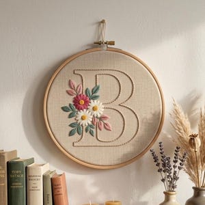 Puede incluir: Un bastidor de bordado circular de madera que contiene tela beige con una letra B grande bordada. La letra está decorada con flores bordadas en tonos rosa, blanco y amarillo, acompañadas de hojas verdes en el lado izquierdo.