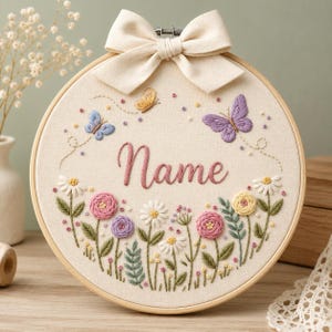 Pode incluir: Um bastidor de bordado circular de madeira contendo tecido cor de creme com desenhos florais bordados à mão, duas borboletas e a palavra Name em letra cursiva rosa. Um grande laço de tecido cor de creme está preso ao topo do bastidor.