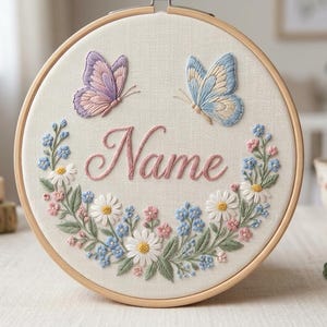 Pode incluir: Um bastidor de bordado circular de madeira contendo tecido branco sujo com a palavra Name bordada em fio rosa cursivo. O design apresenta uma borboleta roxa, uma borboleta azul e uma guirlanda floral de margaridas brancas e pequenas flores azuis.