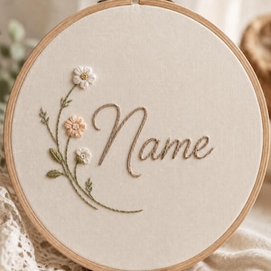 May include: A circular wooden embroidery hoop containing off-white fabric. The fabric features hand-stitched embroidery of the word Name in a brown cursive script, accompanied by a delicate floral stem with white and peach-colored flowers.