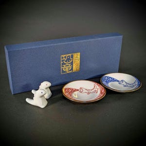 May include: Two white ceramic dinosaur-shaped chopstick rests, one small red and white patterned dish with a dinosaur graphic, and one small blue and white patterned dish with a dinosaur graphic, presented in a blue box with gold Japanese text.