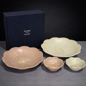 Könnte beinhalten: Ein Set aus vier Keramikschalen mit gewelltem Rand und gerippter Textur, bestehend aus zwei großen und zwei kleinen Schalen in gedeckten Rosa- und Cremetönen mit floralen Details in der Mitte, präsentiert mit einer dunkelblauen Box mit M-mode Aufschrift.