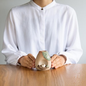 Może przedstawiać: Ręcznie wykonany ceramiczny wazon o unikalnym designie. Wazon ma naturalną paletę kolorów z motywem ptaka i gniazda. Wazon ma mały otwór u góry oraz dekoracyjnego ptaka i gniazdo z przodu. Wazon jest trzymany przez osobę.