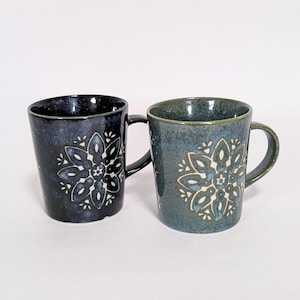 May include: Two ceramic mugs with floral designs. One mug is dark blue with a black handle, the other is teal with a teal handle. Both mugs have a white and blue floral pattern. The mugs are on a white background.
