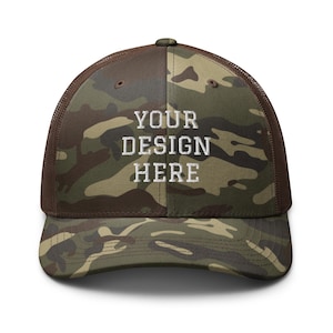 May include: A camouflage print trucker hat with brown mesh back panels and a curved brim. The front panel features the white embroidered text "YOUR DESIGN HERE" centered in a bold, block-style font across the crown of the cap.