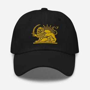 Puede incluir: Gorra de béisbol negra con un diseño dorado bordado que presenta un león sosteniendo una espada frente a un sol. La gorra tiene una visera curva y una correa ajustable.