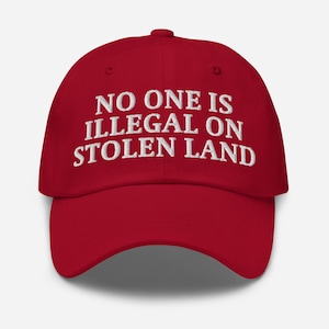 May include: A red baseball cap featuring the embroidered phrase "NO ONE IS ILLEGAL ON STOLEN LAND" in white. The cap has a curved peak and an adjustable strap at the back.