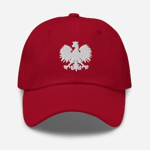 Puede incluir: Una gorra de béisbol de color rojo intenso con un emblema de águila bordado en blanco centrado en el panel frontal. La gorra tiene una corona estructurada, una visera curva y un botón en la parte superior, mostrando un diseño limpio con un águila.