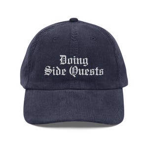 Könnte beinhalten: Eine Baseballkappe aus marineblauem Cord mit dem in Weiß gestickten Text Doing Side Quests in einer gotischen Schriftart. Die Kappe hat eine strukturierte Krone, einen gebogenen Schirm und einen Knopf in der Mitte der Oberseite.