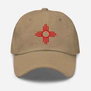 May include: A beige baseball cap with a red embroidered Zia symbol, a Native American sun symbol. The cap has a curved peak and an adjustable strap. The Zia symbol is in the centre of the cap.
