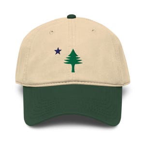 May include: A beige baseball cap with a dark green brim and a small dark green button on top. The front features an embroidered design of a green pine tree positioned to the right of a small, five-pointed dark blue star.