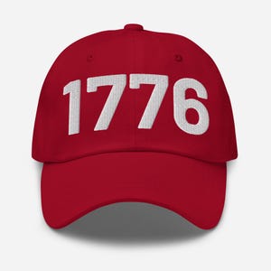 Könnte beinhalten: Rote Baseballkappe mit der in Weiß gestickten Jahreszahl "1776" auf der Vorderseite. Die Kappe hat einen gebogenen Schirm und einen verstellbaren Riemen.