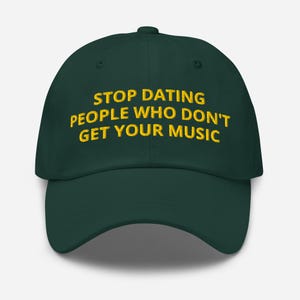 May include: A dark green baseball cap with the embroidered text "STOP DATING PEOPLE WHO DON'T GET YOUR MUSIC" in yellow. The cap has a curved brim and a button on top.