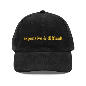 Puede incluir: Una gorra de béisbol de pana negra con el texto expensive & difficult bordado en hilo amarillo en el panel frontal. La gorra tiene una corona estructurada, una visera curva y un botón en la parte superior de la estructura de seis paneles.
