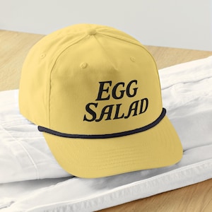 Könnte beinhalten: Gelbe Baseballkappe mit dem schwarzen Schriftzug "EGG SALAD". Die Kappe hat eine schwarze Kordel am Rand und einen gebogenen Schirm. Der Hut liegt auf einer weißen Oberfläche.