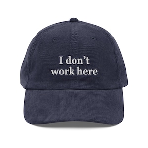 以下が含まれることがあります： 白い文字で「I don't work here」と刺繍された、ネイビーブルーのコーデュロイベースボールキャップ。カーブしたつばとトップにボタンが付いています。カジュアルな普段使いのアクセサリー。