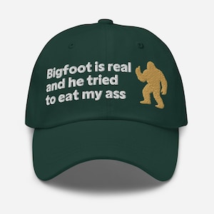 Może przedstawiać: Ciemnozielona czapka z daszkiem z białym haftowanym napisem, Bigfoot is real and he tried to eat my ass, oraz złotym haftowanym sylwetką stojącej postaci Bigfoot umieszczoną po prawej stronie przedniego panelu czapki.