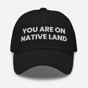 May include: Black baseball cap with the white embroidered text "YOU ARE ON NATIVE LAND". The cap has a curved brim and an adjustable strap at the back. The hat is a classic, casual accessory.