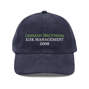 Op de afbeelding: Marineblauwe corduroy baseballcap met de tekst "LEHMAN BROTHERS RISK MANAGEMENT 2008" in groen en wit. De cap heeft een gebogen klep en een ronde bovenkant.