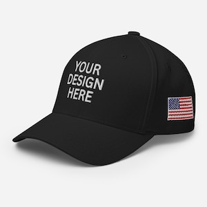 May include: A black baseball cap featuring the white embroidered text YOUR DESIGN HERE on the front panel. The side of the cap displays a small, rectangular embroidered patch of the United States flag with red, white, and blue horizontal stripes.