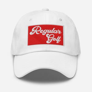 Op de afbeelding: Witte baseballcap met een gebogen klep. De cap heeft de geborduurde tekst "Regular Golf" in wit script op een rode rechthoekige patch. De hoed is gemaakt van een zachte stof en heeft een verstelbare band.