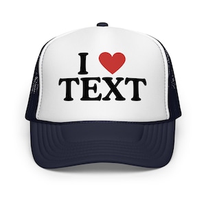 Könnte beinhalten: Eine dunkelblaue und weiße Trucker-Cap mit einer weißen Vorderseite, auf der der schwarze Text I, ein rotes Herzsymbol und der schwarze Text TEXT in einer fetten Serifenschrift gedruckt sind. Die Cap hat einen dunkelblauen Schirm und ein Netz.