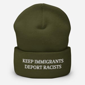 May include: Olive green beanie with the embroidered text "KEEP IMMIGRANTS DEPORT RACISTS" in white. The beanie is a soft, knitted hat, suitable for cold weather. The hat is a solid color with a folded brim.