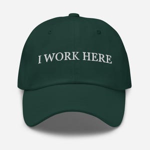 Op de afbeelding: Een donker bosgroene baseballpet met de tekst I WORK HERE geborduurd in witte hoofdletters op het voorpaneel. De pet heeft een gestructureerde kroon, een gebogen klep en een knoopje bovenop het midden van de bol.