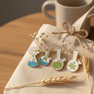 Puede incluir: Cuatro pines de esmalte con forma de matraces de laboratorio, cada uno con un diseño diferente. Uno presenta margaritas, otro rosas, un tercero una planta verde y el último una hoja de cannabis verde. Los pines son blancos, azules y verdes.