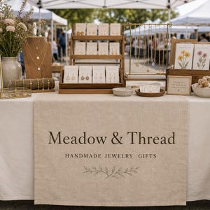 Op de afbeelding: Een sieradendisplaytafel met een beige linnen loper met de tekst Meadow & Thread HANDMADE JEWELRY GIFTS. De display bevat houten oorbellenstandaards, een gouden kettingrek, een houten buste met gelaagde kettingen en bloemenkaarten.