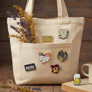 May include: A cream-colored tote bag with a front pocket, adorned with various enamel pins. The pins feature designs like a gold ticket, a beer mug, and quotes. The bag also holds dried lavender and yellow flowers.
