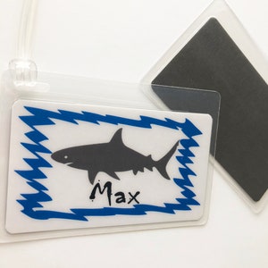 May include: A white luggage tag with a blue and white shark design. The tag has the name "Max" written on it.
