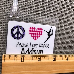 May include: A white luggage tag with a clear plastic loop. The tag features a peace sign, a checkered heart, and a dancer silhouette. Text on the tag reads "Peace Love Dance" and "Addisun". A wooden ruler is placed below the tag.
