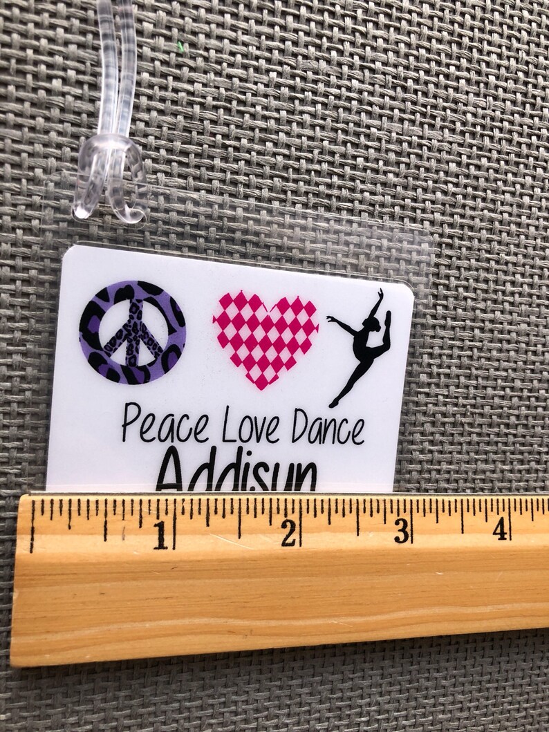 May include: A white luggage tag with a clear plastic loop. The tag features a peace sign, a checkered heart, and a dancer silhouette. Text on the tag reads "Peace Love Dance" and "Addisun". A wooden ruler is placed below the tag.
