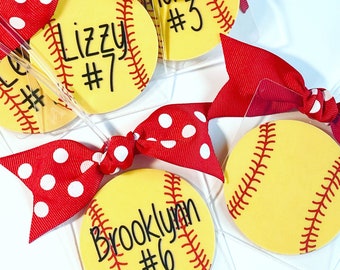 May include: Six softball-themed keychains with red and white polka dot bows. Each keychain features a yellow softball with black stitching and a name and number. The names are "Lizzy #7", "Brooklyn #6", "Riley #3", and three others.