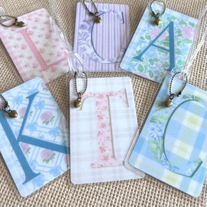 May include: A collection of rectangular luggage tags featuring various letters of the alphabet, including L, C, A, K, T, and G. Each tag is encased in clear plastic and includes a small bronze-colored heart charm attached by a ball chain.