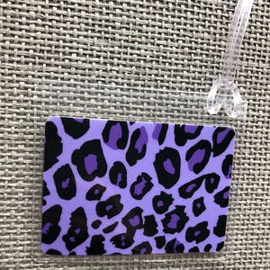 May include: A rectangular luggage tag with a purple leopard print design and a clear plastic loop for attachment. The tag features black spots on a light purple background.