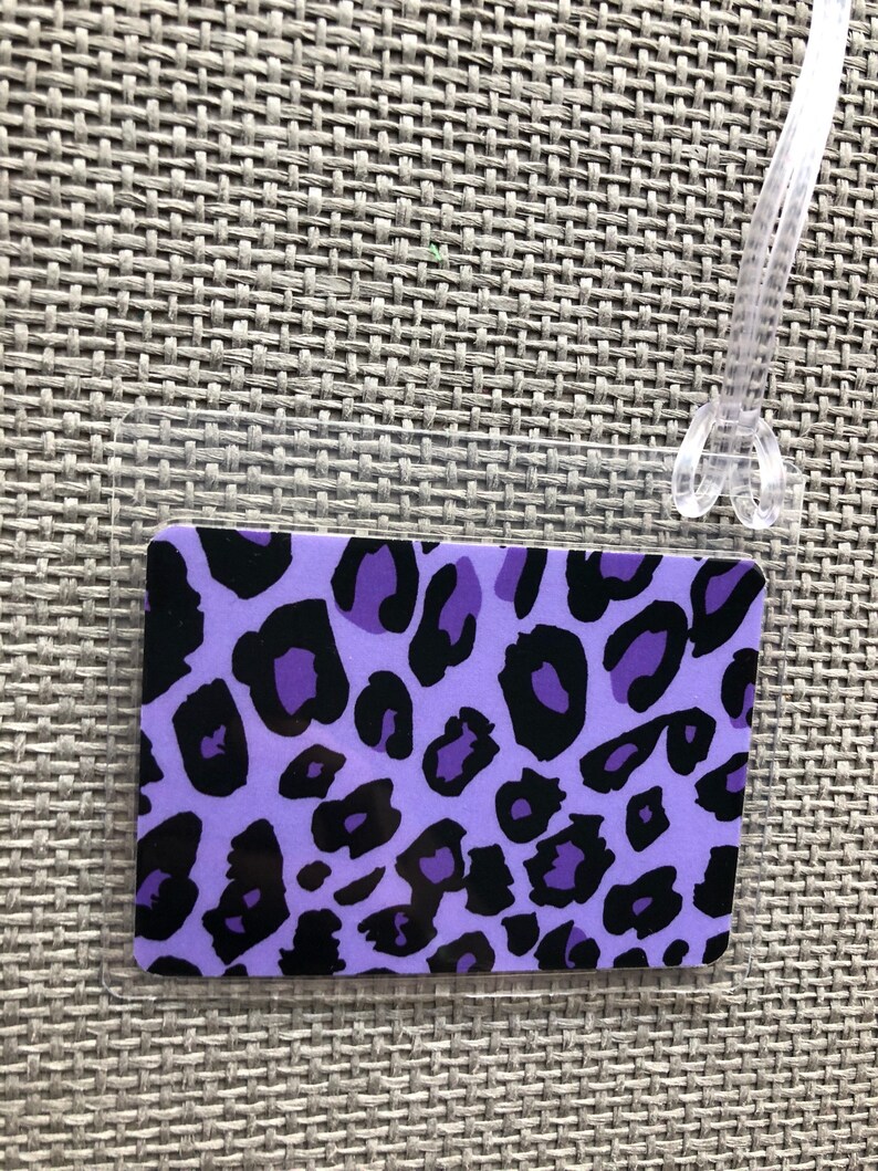 May include: A rectangular luggage tag with a purple leopard print design and a clear plastic loop for attachment. The tag features black spots on a light purple background.
