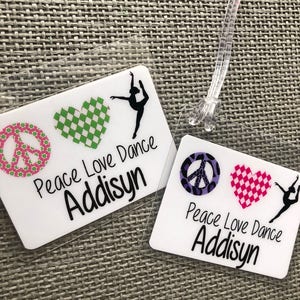 May include: Two clear luggage tags with the text "Peace Love Dance Addisyn". One tag features a green checkered heart and a peace sign. The other tag has a pink checkered heart and a peace sign.