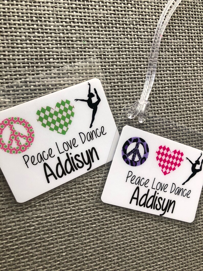 May include: Two clear luggage tags with the text "Peace Love Dance Addisyn". One tag features a green checkered heart and a peace sign. The other tag has a pink checkered heart and a peace sign.