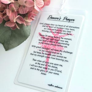 May include: A clear plastic tag with the text "Dancer's Prayer" and a pink ballerina graphic. The tag is next to pink flowers.