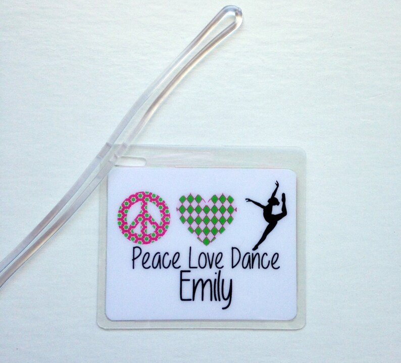 May include: A white luggage tag with a clear plastic loop. The tag features a peace sign with a pink and green floral pattern, a pink and green checkered heart, and a black silhouette of a dancer. The words "Peace Love Dance Emily" are printed in black.