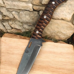 Puede incluir: Un cuchillo con mango marr&oacute;n y negro con un acento de metal plateado en el mango. El cuchillo tiene una hoja negra y est&aacute; sobre una tabla de cortar de madera.