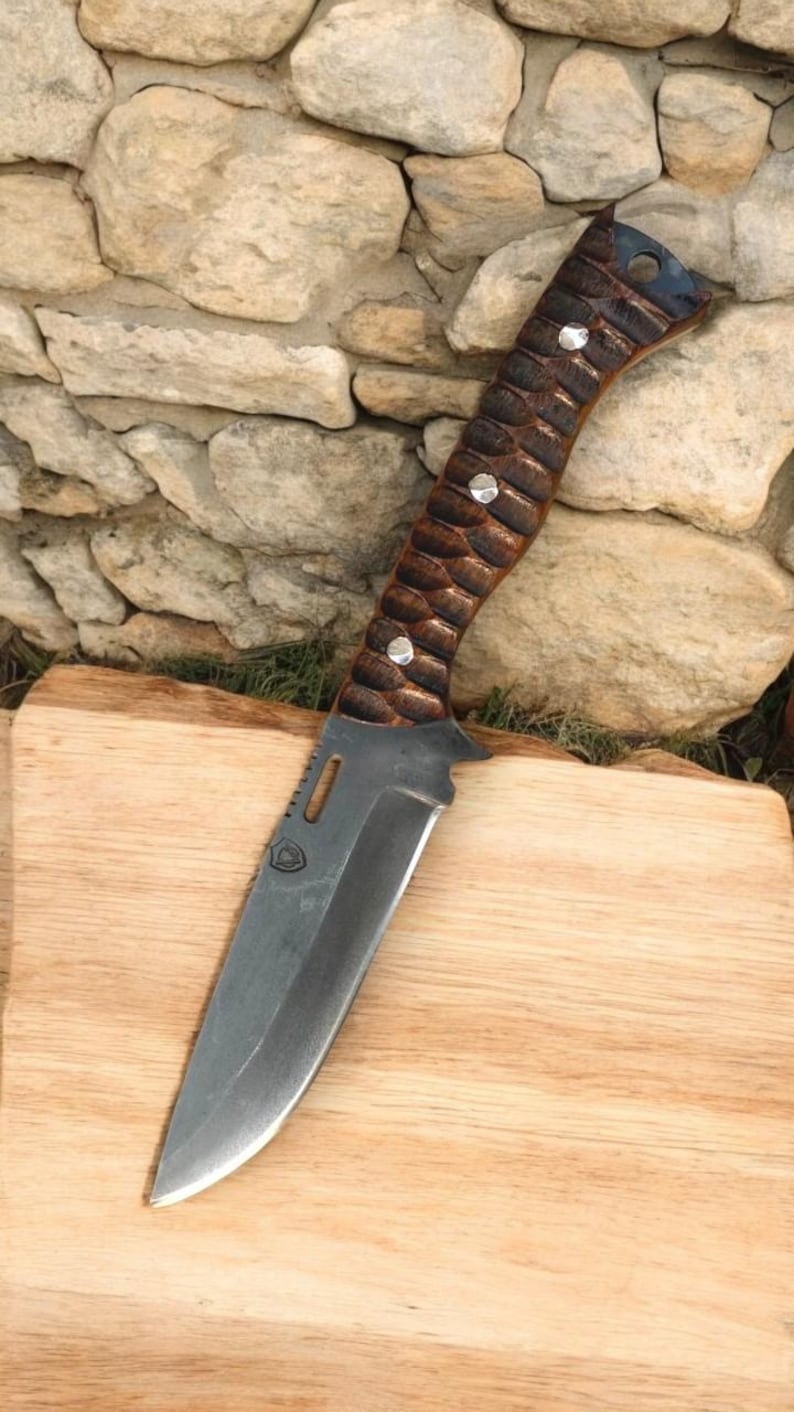 Puede incluir: Un cuchillo con mango marr&oacute;n y negro con un acento de metal plateado en el mango. El cuchillo tiene una hoja negra y est&aacute; sobre una tabla de cortar de madera.
